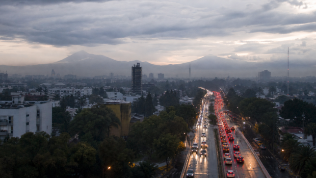Tráfico vehicular en una avenida de la Ciudad de México al amanecer, con pavimento mojado y cielo nublado.